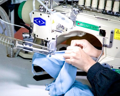 A close-up of a skilled technician using a professional flatlock sewing machine to create durable, smooth seams on light blue athletic apparel.